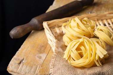 Çiğ tagliatelle yuvalarının üst görüntüsü çuval, sepet, mutfak oklavası ve kırsal ahşap masa, siyah arkaplan, yatay