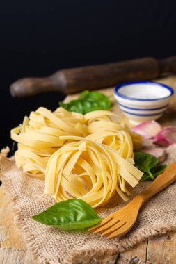 Çiğ tagliatelle yuvalarının üst görüntüsü, fesleğen, sarımsak ve mutfak merdanesi çuval ve kırsal ahşap masa, siyah arkaplan, dikey