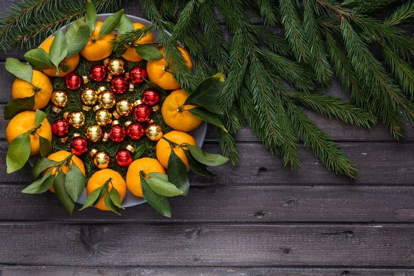 Mandarines de Noël avec des feuilles se trouvent dans une assiette couverte d'aiguilles d'un arbre de Noël et de jouets de Noël — Photo