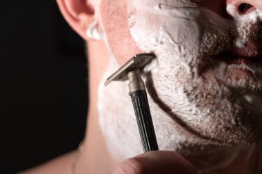 Bir adam yanağını keskin olmayan bir bıçakla tehlikeli bir aletle tıraş eder..