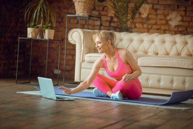 Genç formda blogcu kız spor kıyafetleriyle yoga minderinde oturuyor evde online antrenman yapıyor, evden çevrimiçi aerobik dersleri kaydediyor, kamera üzerinde egzersiz yapıyor.