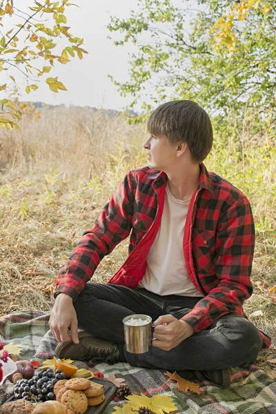 Autumn picnic in the park, warm autumn day. The guy is holding a cup of cocoa. Warm cocoa with marshmallows in a metal mug. Autumn concept.