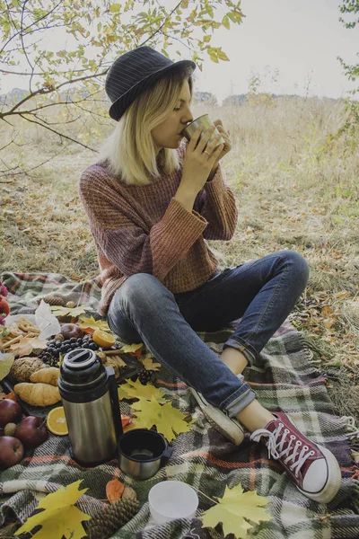 Autumn picnic in the park, warm autumn day. The girl holds a cup with tea in her hands. Basket with flowers on a blanket in yellow autumn leaves. Autumn concept.
