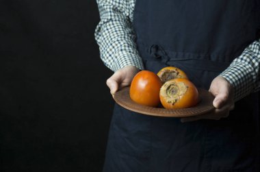 Aşçı elinde bir hurma tutuyordu. Erkek eli taze hurma dolu bir tabak tutar. Mevsimlik meyveler. Karanlık arka plan. Metin için yer.