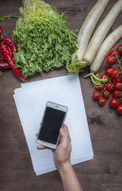 Bir adam elindeki akıllı telefonu mutfak masasında taze sebzelerin arka planına karşı tutar. Yemek arkaplanı. Daire yalan söylüyordu. Mesaj için yer. Arka plan menü masası. Sağlıklı denge 