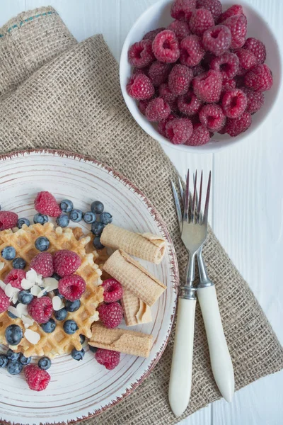 Waffles with fresh banana, raspberries, blueberries for breakfast. Fresh waffles with fruits. Light wooden background. Summer homemade breakfast. Copy space. The balance of healthy eating.
