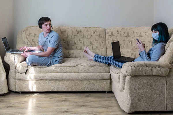 A young family works from home on self-isolation. A man sits on the edge of the couch and works on a laptop with a headset. He turned to a masked woman using the phone. Keeping distance quarantined.