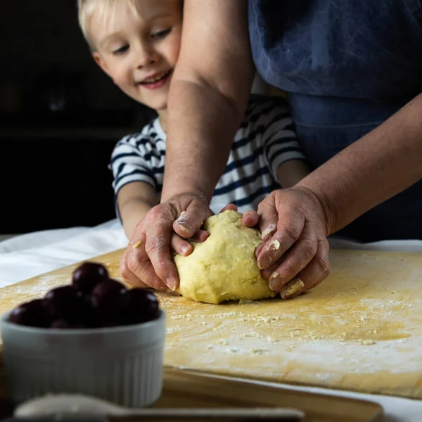A child with the help of grandmother's hands kneads the dough on a wooden table in flour. Tender care for a new generation, teach cooking. Joint family pastime on self-isolation.