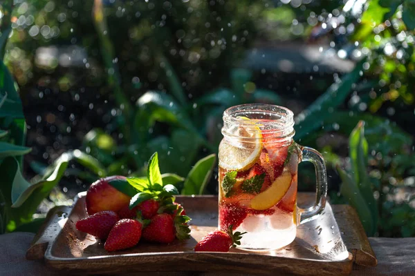 Fruit and berry homemade lemonade from strawberries, peach, lemon. Cold carbonated drink in a glass jar on wooden tray with fruits. On backdrop the green grass in the garden are soaring bokeh.