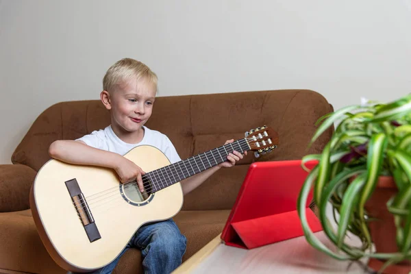 Cute blond boy plays the guitar and looks at the tablet standing on the table. A fun online lesson on the internet. Digital education in self isolation mode. Modern lifestyle. Stay at home.