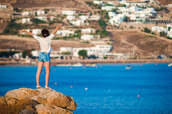 Giovane ragazza felice sul bordo di una scogliera con splendida vista sul vecchio piccolo villaggio di Mykonos, in Grecia — Foto Stock