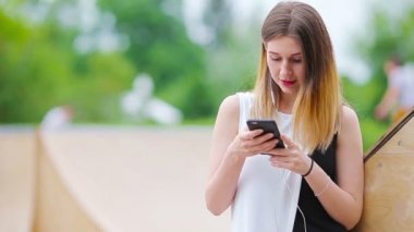 Beyaz bir kadın mesaj gönderiyor ve Avrupa 'da açık havada müzik dinliyor. Güneş gözlüklü güzel kız ahşap bankta akıllı telefon kullanarak oturuyor.