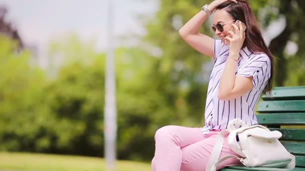 Joven chica caucásica hablando por su teléfono inteligente al aire libre. Hermosa chica en gafas de sol en la calle utilizando el teléfono inteligente y hablando por teléfono con su amigo — Vídeo de stock