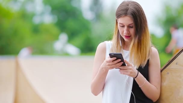 Νεαρή καυκάσια γυναίκα στέλνει μήνυμα και ακούει μουσική σε εξωτερικό χώρο στην ευρωπαϊκή πόλη. Όμορφο κορίτσι με γυαλιά ηλίου κάθεται σε ξύλινο πάγκο χρησιμοποιώντας smartphone — Αρχείο Βίντεο