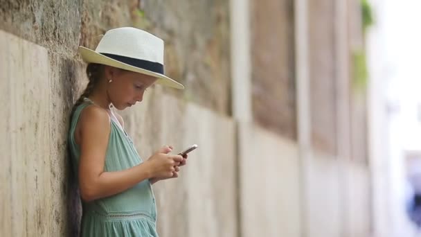 Rozkošná holčička s chytrým telefonem v teplý den venku v evropském městě — Stock video