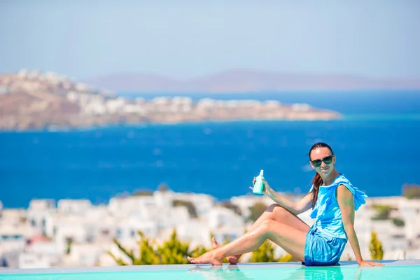 Young woman applying sunscreen on her legs, sitting on the edge of pool background old town Mykonos in Europe — Stock Photo, Image