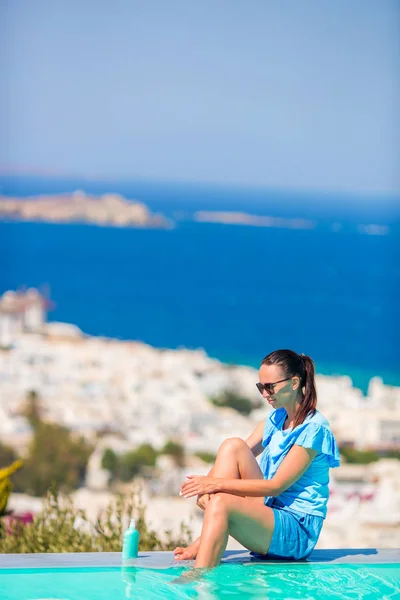 Mulher magro aplicando protetor solar em suas pernas, sentado na borda da piscina fundo cidade velha Mykonos na Europa — Fotografia de Stock