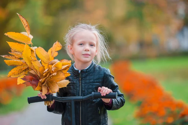 Portrait of adorable little girl with yellow leaves bouquet in fall on scooter — Stock Photo, Image