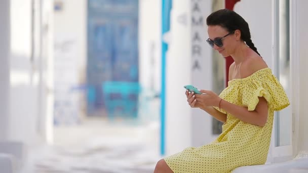 Krásná žena s chytrý telefon venku v starých ulic Mykonu. Dívka v ulici typické řecké tradiční vesnice s bílými stěnami a barevnými dveřmi na ostrově Mykonos, Řecko, Evropa — Stock video