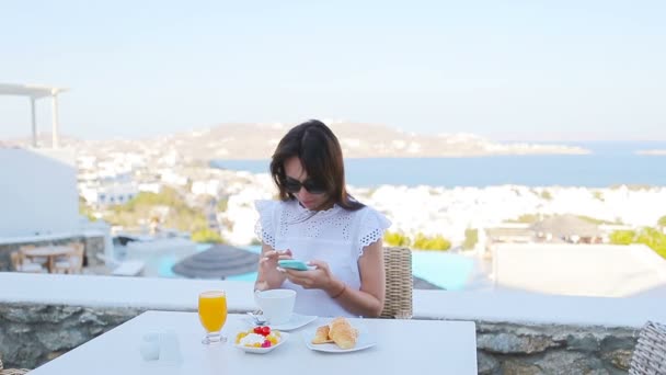 Mooie elegante dame ontbijten op terras met geweldig uitzicht op Mykonos stad. Warme koffie drinken op het terras van luxe hotel met uitzicht op zee in resort restaurant vrouw. — Stockvideo