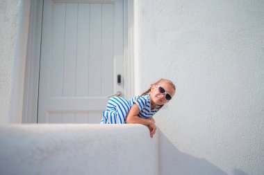 Sevimli kız açık havada eski sokaklar bir Mykonos. Beyaz duvarları ve Yunanistan'da Mykonos Adası renkli kapı ile tipik Yunan geleneksel köy Street çocuk