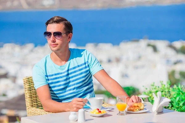 Niño desayunando en la cafetería al aire libre con una vista increíble de la ciudad de Mykonos. Hombre bebiendo café caliente en la terraza del hotel de lujo con vistas al mar en el restaurante del complejo . — Foto de Stock
