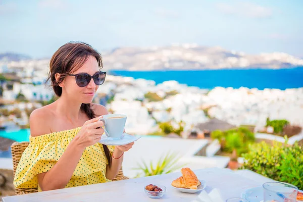 Menina bonita tomando café da manhã no café ao ar livre com vista incrível sobre a cidade de Mykonos. Mulher bebendo café quente no terraço do hotel de luxo com vista para o mar no restaurante resort . — Fotografia de Stock
