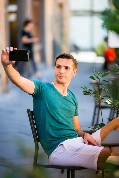 Turista caucásico con teléfono inteligente tomando selfie sentado en la cafetería al aire libre. Joven chico urbano de vacaciones explorando la ciudad europea — Foto de Stock