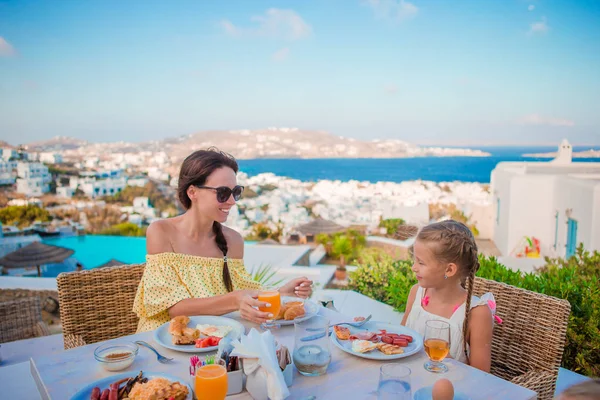 Familia desayunando en la cafetería al aire libre con una vista increíble de la ciudad de Mykonos. Adorable chica y mamá beber jugo fresco y comer croissant en la terraza del hotel de lujo —  Fotos de Stock