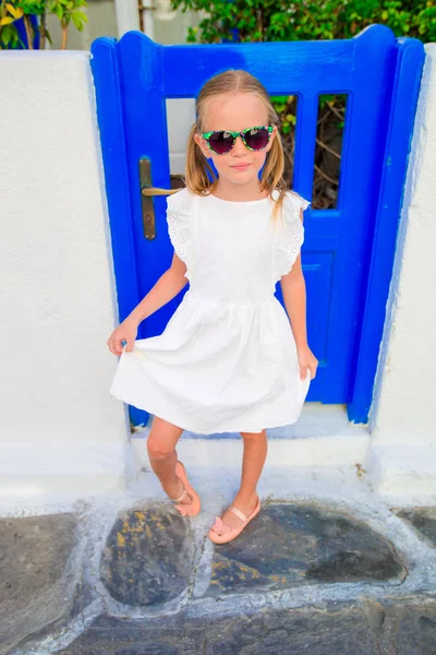 Liten söt tjej porträtt utomhus i gammal grekisk by. Kid på gatan av typisk grekisk traditionell by med vita väggar och färgglada dörrar på Mykonos, Greklands ö — Stockfoto