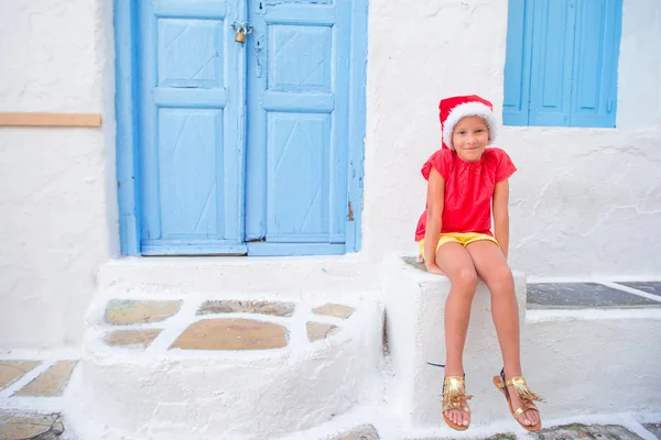 Little girl in santa hat outdoors in old street on Christmas vacation in Mykonos. Kid at street of typical greek traditional village with white walls and colorful doors on Mykonos Island, in Greece — Stock Photo, Image