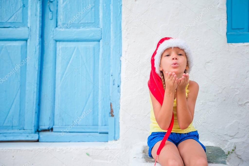 Little girl in santa hat outdoors in old street on Christmas vacation in Mykonos. Kid at street of typical greek traditional village sends kisses