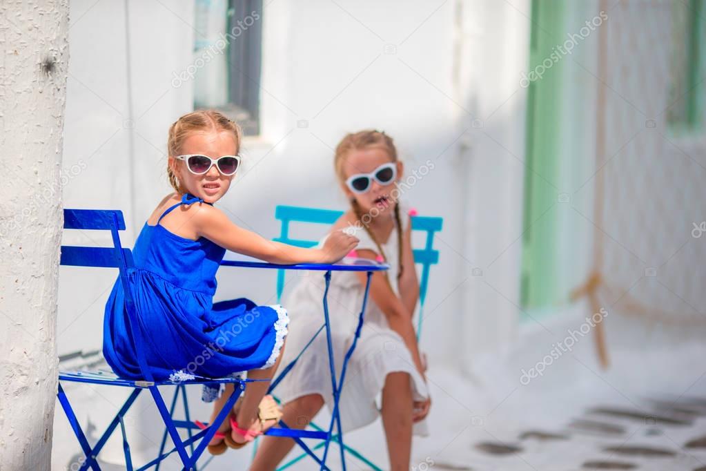 Two girls in blue dresses sitting on blue chairs and table on street of typical greek traditional village with white houses on Mykonos Island, Greece, Europe.