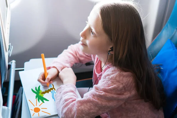 En söt liten flicka som reser med ett flygplan. Kid ritning bild med färgglada pennor sitter nära flygplansfönstret — Stockfoto