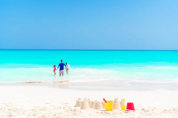 Песчаный замок на белом пляже с пластиковыми детскими игрушками и семьей на морском фоне — стоковое фото