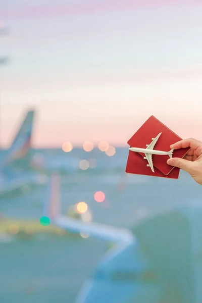 Primo piano mano in possesso di un giocattolo modello di aereo e passaporti presso lo sfondo dell'aeroporto grande finestra — Foto Stock