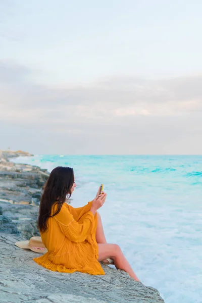 Mooie jongedame met mobiele telefoon in de zonsondergang. Gelukkig meisje in jurk in de avond op het strand — Stockfoto