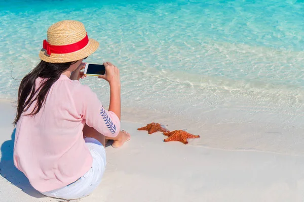 Mujer con teléfono móvil tomar una foto de estrellas de mar rojas en la playa tropical — Foto de Stock