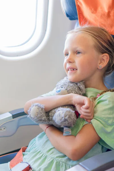 Entzückendes kleines Mädchen, das mit dem Flugzeug reist. Kind sitzt mit seinem Lieblingsspielzeug am Flugzeugfenster — Stockfoto