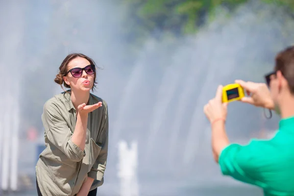 Jeune homme prenant une photo de sa petite amie tout en étant assis derrière la fontaine. Jeune homme faisant la photo de la femme dans la rue riant et s'amusant en été . — Photo