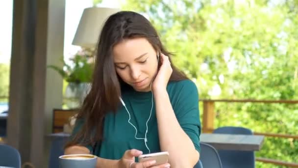 Mujer joven con teléfono celular mientras está sentada sola en la cafetería durante el día caliente. Atractiva hembra con linda sonrisa mensaje de escritura con teléfono móvil mientras descansa en la cafetería — Vídeo de stock