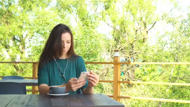 Женщина со смартфоном в кафе пьет кофе, улыбается и пишет смс на мобильный телефон. Портрет красивой девушки — стоковое видео