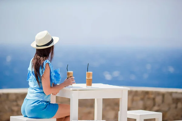 Jóvenes en la cafetería al aire libre disfrutando de vistas al mar. Hermosa mujer relajarse durante vacaciones exóticas en la playa disfrutando de frappe — Foto de Stock