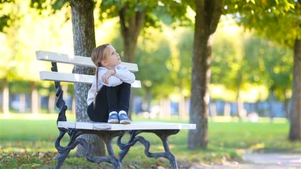 Adorable petite fille à la belle journée d'automne en plein air. Enfant mignon assis sur le banc dans le parc extérieur — Video