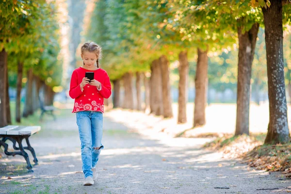 Lilla bedårande flickan med smartphone i höst. Kid har kul på varma soliga höstdag utomhus — Stockfoto