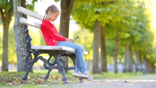 Маленька чарівна дівчинка зі смартфоном восени. Малюк розважається в теплий сонячний осінній день на відкритому повітрі — стокове відео