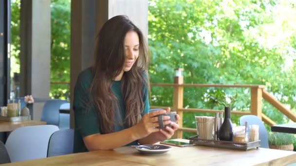 Mujer joven sentada en un café al aire libre bebiendo café. Retrato de chica feliz con taza de café — Vídeos de Stock