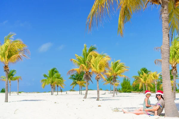 Joven mamá y niña se divierten en la playa tropical en sombrero de santa sentado cerca de la palmera —  Fotos de Stock