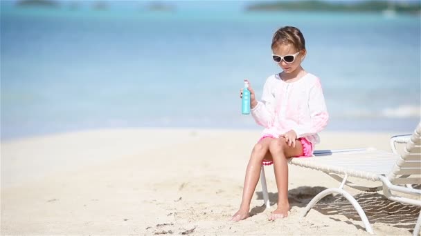 Маленькая очаровательная девочка с бутылкой крема для загара сидит на тропическом пляже — стоковое видео