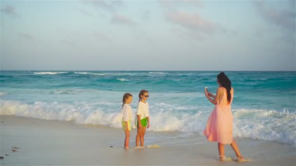 小可爱的女孩和年轻的母亲在热带海滩在温暖的晚上。妈妈在日落时拍一张漂亮的孩子的照片 — 图库视频影像